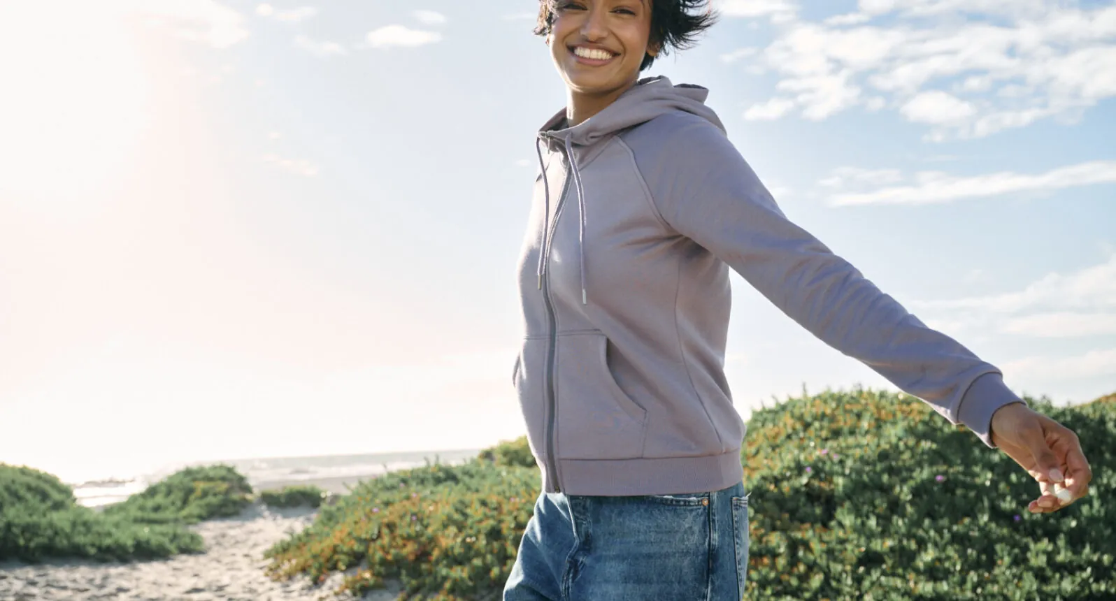 Woman smiling outdoors in a lavender eco-friendly zip-up hoodie, walking along a coastal path.