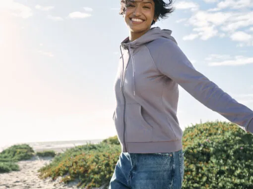 Woman smiling outdoors in a lavender eco-friendly zip-up hoodie, walking along a coastal path.
